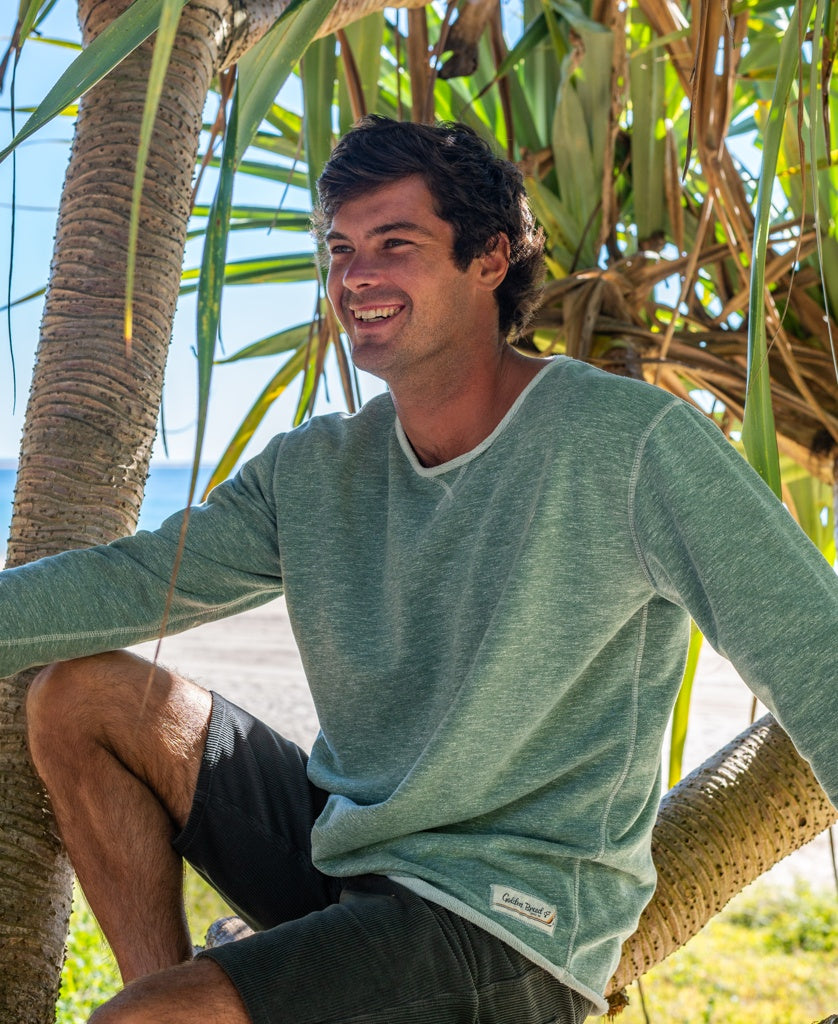 A man wearing the Golden Breed Classic Rag Top in Surf Green Marle and black shorts sits on a tree branch at the beach, smiling with sunlight and palm leaves behind him for an effortless surf look.