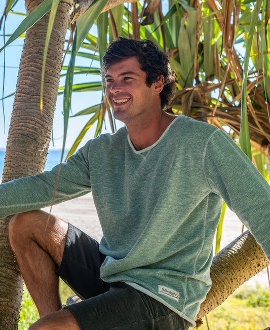 A man wearing the Golden Breed Classic Rag Top in Surf Green Marle and black shorts sits on a tree branch at the beach, smiling with sunlight and palm leaves behind him for an effortless surf look.