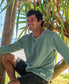 A man wearing the Golden Breed Classic Rag Top in Surf Green Marle and black shorts sits on a tree branch at the beach, smiling with sunlight and palm leaves behind him for an effortless surf look.