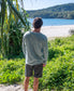 Wearing a Golden Breed Classic Rag Top in Surf Green Marle and shorts, a person stands on grass gazing at the sandy beach and ocean with hills beyond, embodying an effortless surf style.