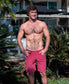 A shirtless man stands outdoors in front of palm plants and a modern building, wearing Golden Breeds Floyd Cord Walkshort in Scarlet with his hands in his pockets.