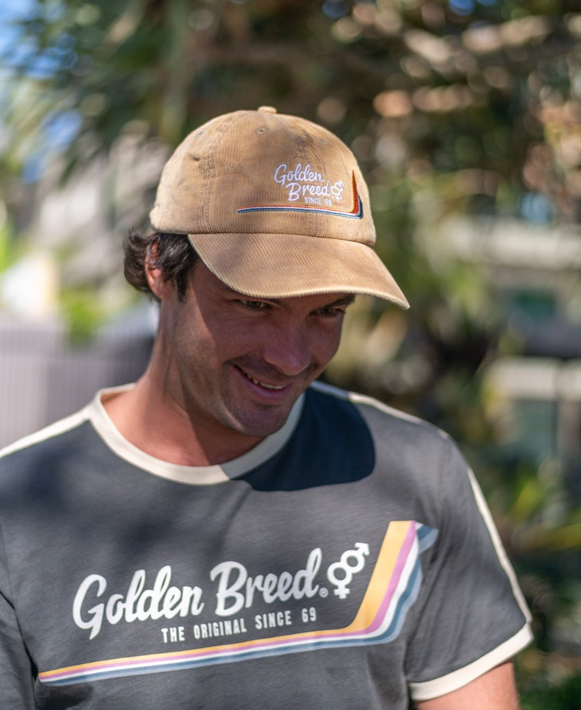 A man wearing a Golden Breed Hancock Cord Cap in camel and a matching T-shirt stands outdoors, looking down and smiling in the sunlight, highlighting the classic unisex adjustable hat style.