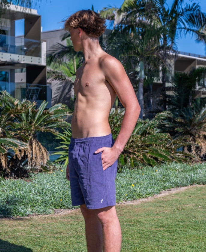 A shirtless person wearing Golden Breeds Floyd Cord Elastic Walkshort in Port stands on grass near palm trees, looking to the left with modern buildings visible in the background.