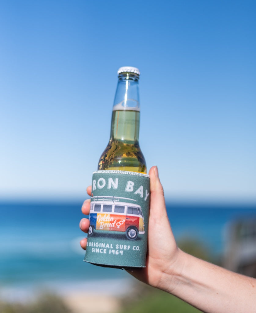 A hand holds a Byron Stubby Kombi | Surf Green by Golden Breed, with ocean and blue sky in the background, capturing the Byron Bay surf vibe.