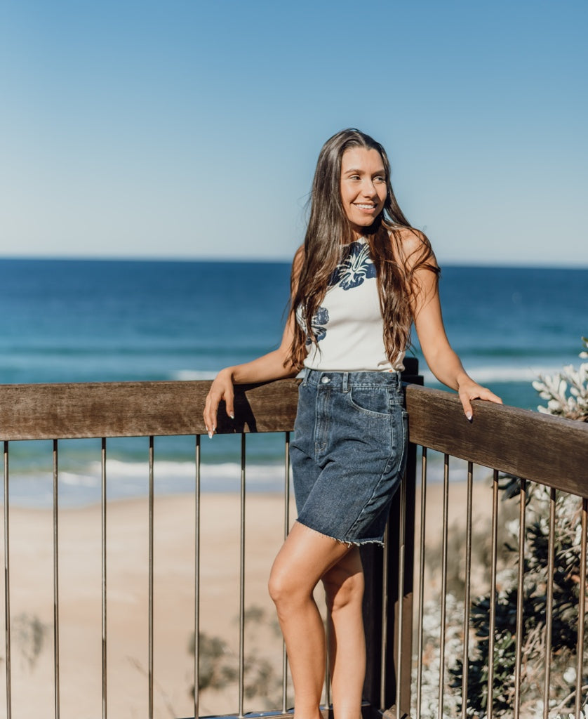 A woman stands on a wooden deck overlooking the ocean, wearing a white top and Golden Breed’s Izzy Short in Dark Blue, with a clear blue sky above and sandy beach below.