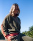 A woman in the Golden Breed Peggy 1/2 Zip Retro sweatshirt in Dk Khaki/Red sits outdoors on a sunny day, with trees and a clear blue sky behind her.