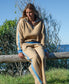 Wearing the Golden Breed Peggy 1/2 Zip Retro in Beige/Azure, a person sits on a wooden fence outdoors, with trees and water in the background.