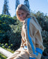 A person with long blonde hair wears the Golden Breed Peggy 1/2 Zip Retro in Beige/Azure, sitting on a wooden rail outdoors surrounded by trees and bushes.