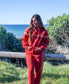 A person in a red Golden Breed Long Island Hoodie stands outdoors by a wooden fence, with greenery and the ocean visible in the background.