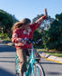 A woman in a Golden Breed Hibiscus Sweat | Wash Red/Beige sweater and brown pants rides a turquoise bike with one hand raised on a sunny, green-lined road.