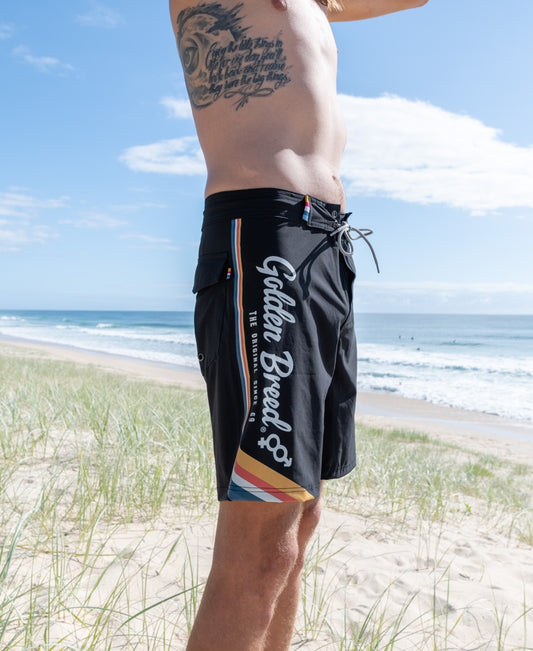 Someone wearing Golden Breeds Retro Surf Boardie in Black stands on a sandy beach with grassy dunes and the ocean behind them.
