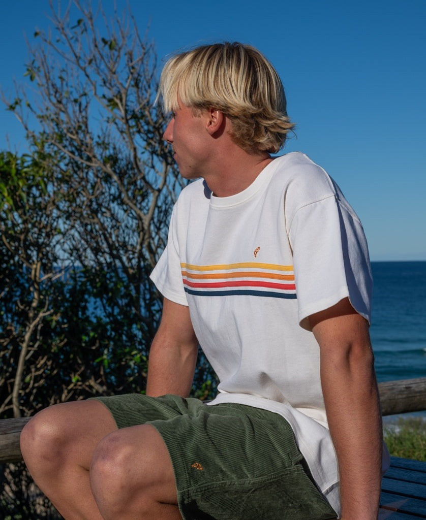 A person with blonde hair sits outdoors on a bench, wearing a white t-shirt with colorful stripes and Golden Breeds Floyd Elastic Waist Walkshort in Khaki, looking toward the ocean and blue sky.