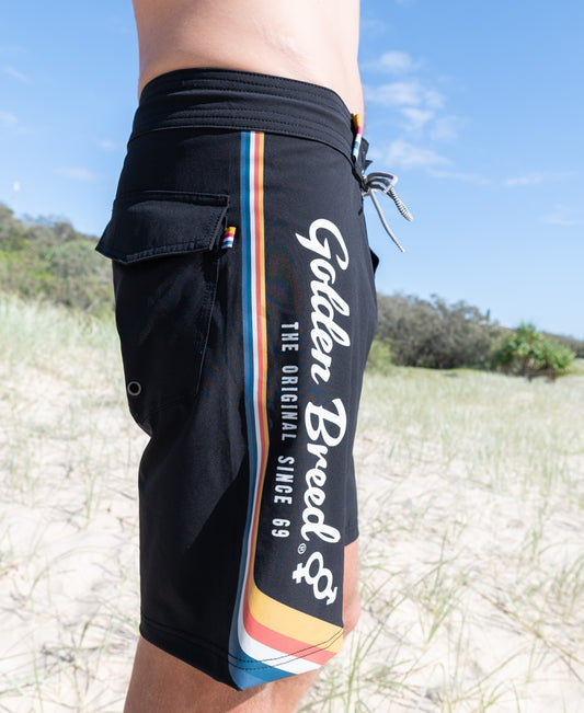 A person stands on a sandy beach in Retro Surf Boardie | Black by Golden Breed, featuring rainbow stripes, The Original Since 69 text, recycled polyester fabric, and 4-way stretch. Grass and trees are in the background.