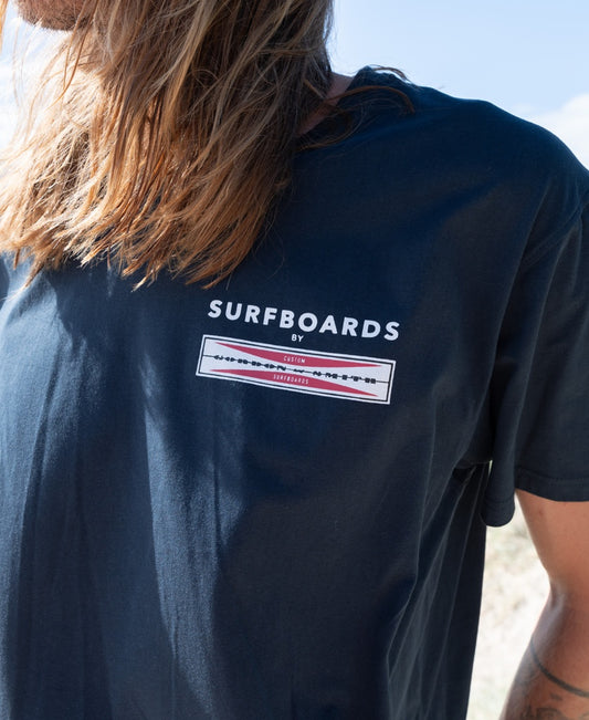A person wears a navy 100% cotton G&S Corp Logo Tee by Gordon & Smith, featuring SURFBOARDS BY and a chest graphic. The face is not visible—a classic pick for surf apparel fans.