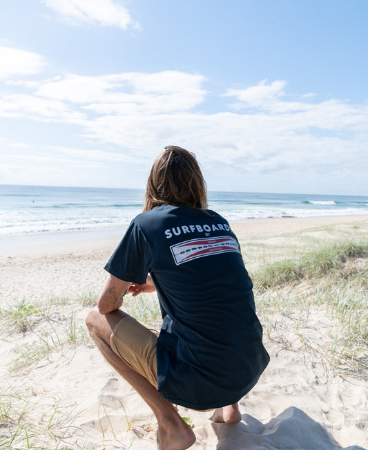 A person with long hair squats on sandy dunes, facing the ocean in a navy G&S Corp Logo Tee by Gordon & Smith and tan shorts. The partly cloudy sky sets the scene for fans of classic surf apparel.