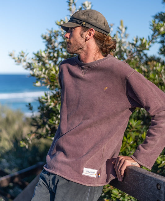 A man with long hair in a cap and the Golden Breed Rev Sweater | Wash Plum stands outdoors by greenery, glancing sideways with the ocean behind him, embodying a relaxed surf vibe.