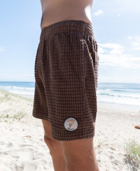 A person wearing Golden Breeds El Shorto Walkshort in Wash Brown, featuring a small round patch and elastic waist, stands on a sandy beach with the ocean and blue sky behind them.