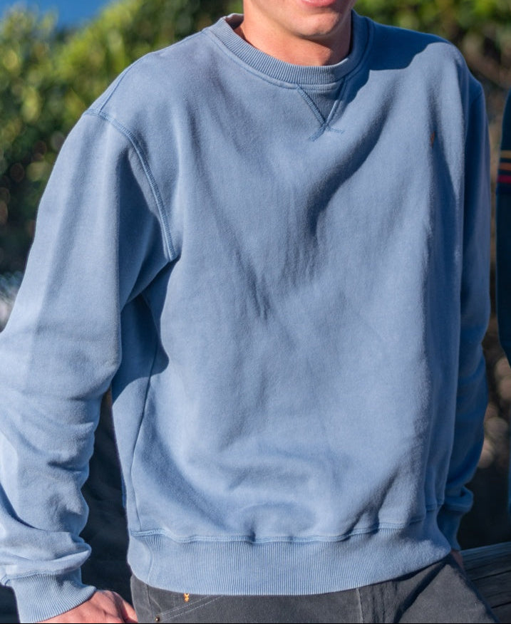 A person wearing the Golden Breed Tide Sweater | Denim, made from cotton brushed fleece, and dark pants stands outdoors with greenery in the background.