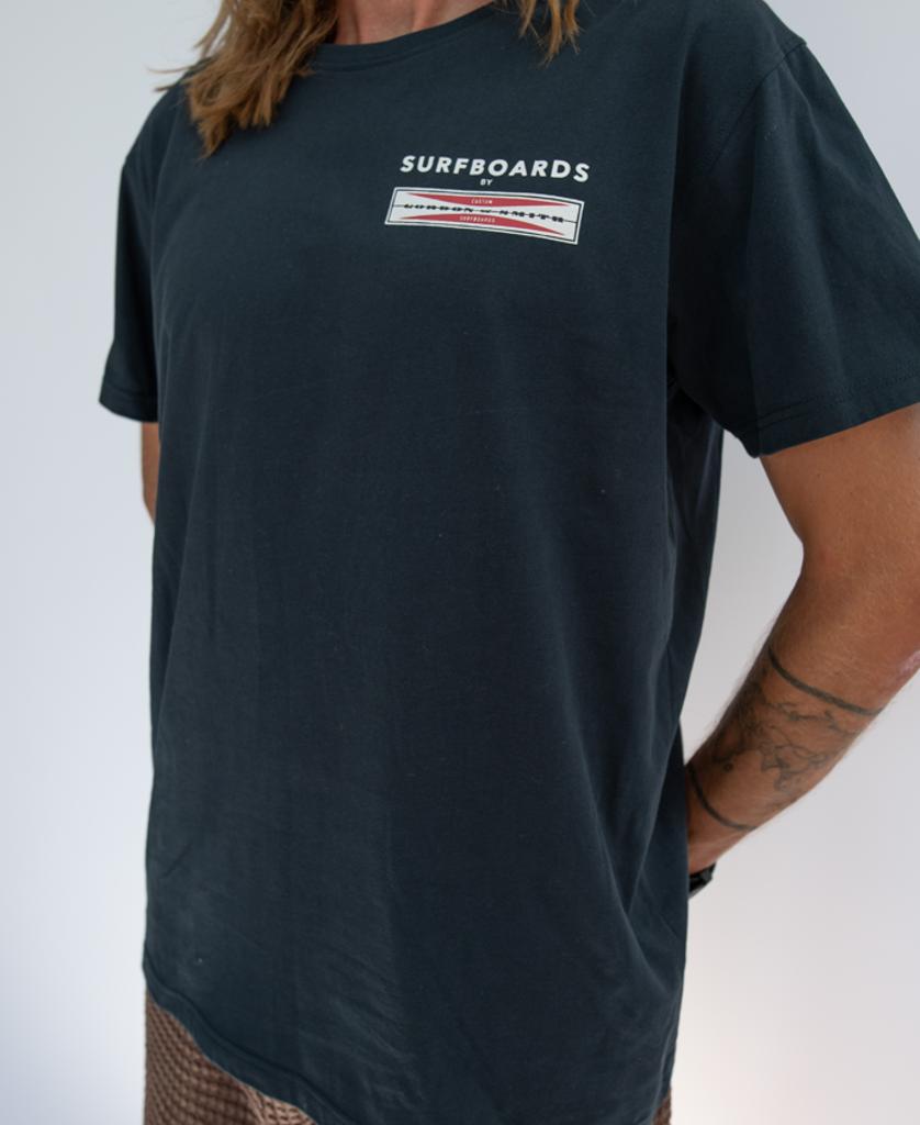 A person stands against a plain white background wearing the navy Gordon & Smith G&S Corp Logo Tee, featuring Surfboards text and chest graphic. Made from 100% cotton surf apparel. Their arms, partially visible, show a tattoo.