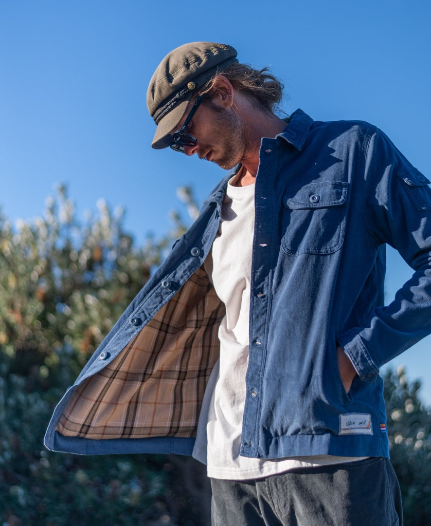 A man stands outdoors under a blue sky, looking down as he opens his shirt to reveal the plaid cord cotton flannel lining of the Golden Breed Cahoots Cord Jacket in Vintage Navy. Shrubs are visible in the background.