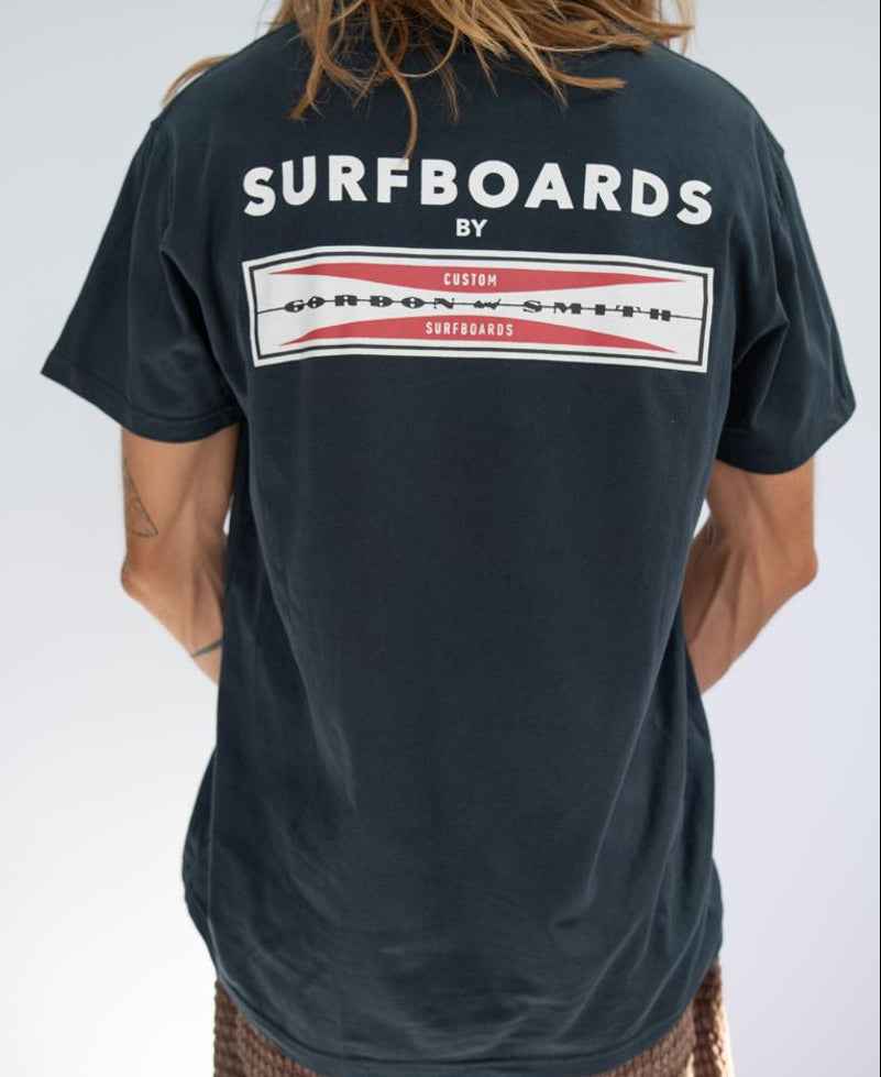 Person wearing a navy G&S Corp Logo Tee by Gordon & Smith, a 100% cotton shirt featuring SURFBOARDS BY GORDON & SMITH CUSTOM SURFBOARDS printed on the back, standing with their back to the camera—a true surf staple.