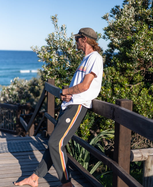 Wearing Trakkydaks | Wash Black by Golden Breed, a person in regular fit casuals and sunglasses leans on a wooden railing overlooking the ocean with greenery nearby.