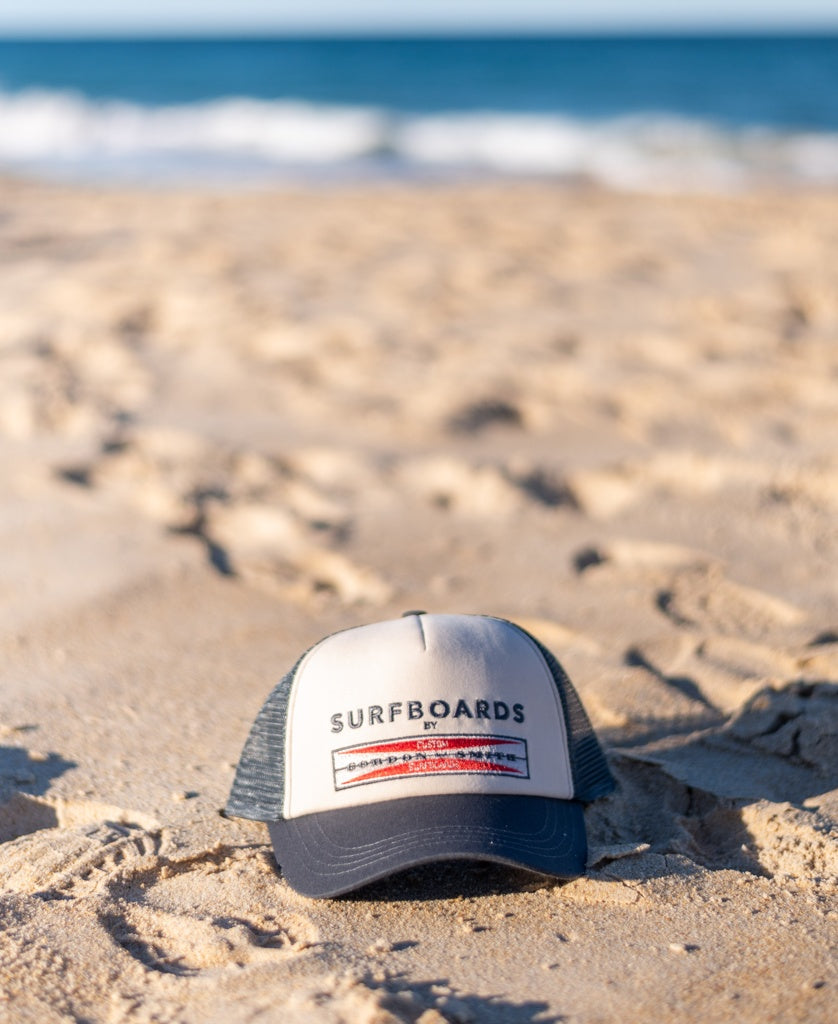 The G&S Boards Trucker Cap in wash black by Gordon & Smith, featuring bold SURFBOARDS logo embroidery, sits on sandy beach with ocean waves in the background.
