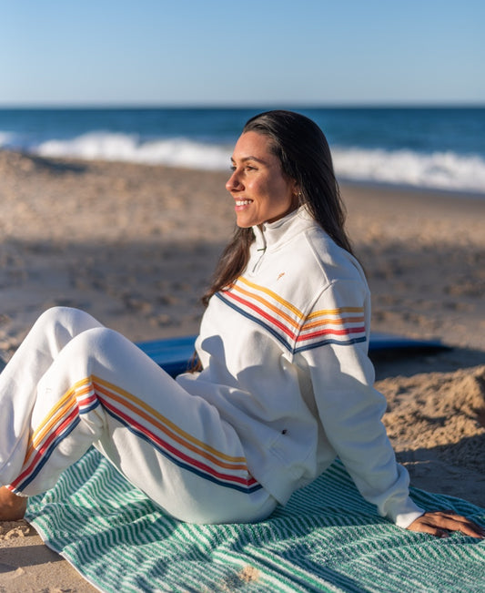 Wearing Golden Breeds Long Island Crunchie T/P in Off White, a woman sits smiling on a green-striped towel at the beach, gazing out at the ocean.