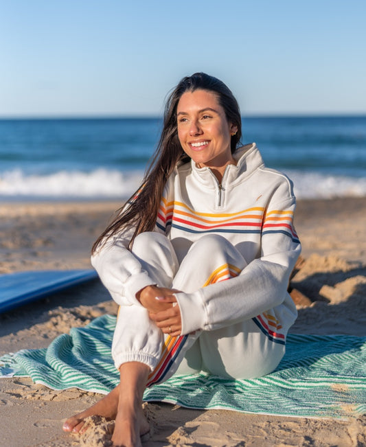 A woman smiles on a striped beach blanket, wearing the Golden Breed Long Island Crunchie T/P in off white. The ocean and blue sky provide a serene backdrop.