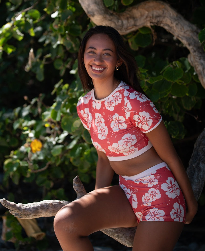 A woman wears the Golden Breed Indie Crop Tee in Hibiscus Wash Red/Beige, sitting on a tree branch outdoors and smiling at the camera, with green leaves in the background.