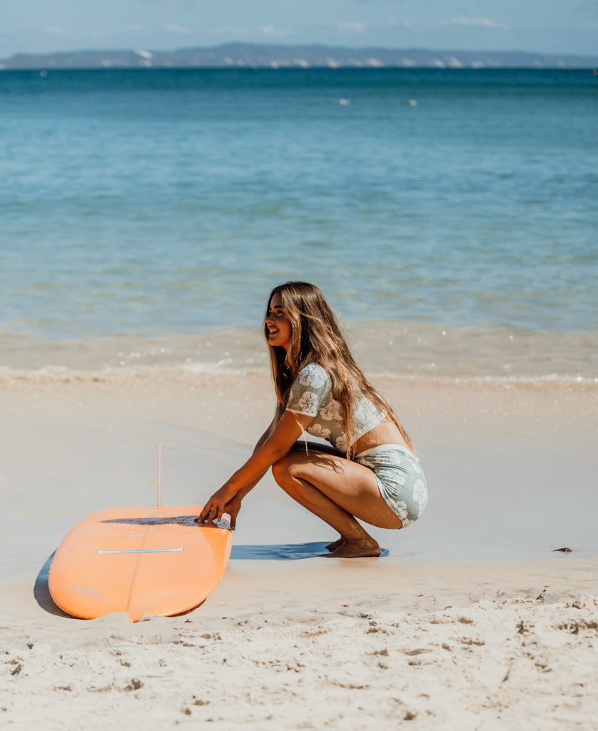 A woman in the Golden Breed Indie Crop Tee in Hibiscus Moss/Off White crouches beside an orange surfboard on a sandy beach with calm blue water in the background.
