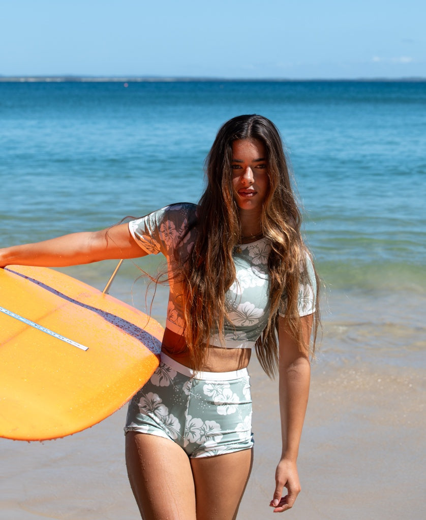 A woman wearing the Golden Breed Indie Crop Tee in Hibiscus Moss / Off White stands on a sandy beach with an orange surfboard, ocean waves, and a blue sky in the background.