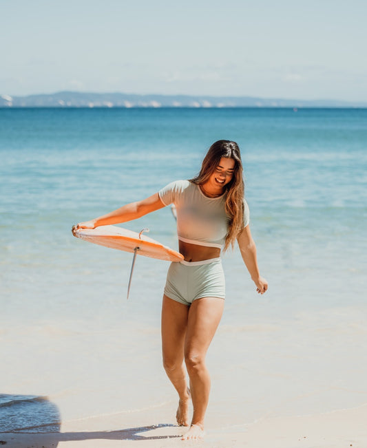 A woman wearing the Golden Breed Indie Crop Tee Retro in Moss/Off White stands on a sandy beach, holding a round orange flotation device near the shoreline.