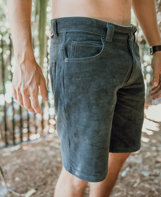 Someone stands outdoors near a fence and trees, wearing Golden Breeds Floyd Cord Walkshort in Wash Black—100% cotton corduroy shorts with a structured waist; their torso is partially out of frame.