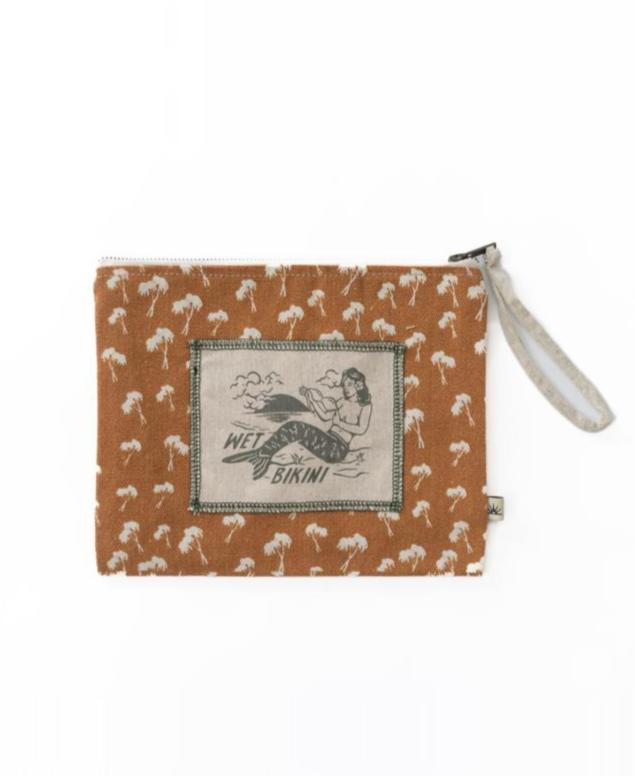 The Bomba WET BIKINI Palm Repeat bag features a brown zippered design with a small white palm tree pattern, waterproof interior, and a label showing a woman with a surfboard—ideal as your go-to beach bag.