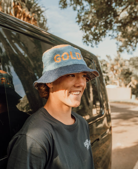 A person in a black T-shirt and the Golden Breed Ferry Terry Bucket Hat in Azure Blue stands outside by a black van, smiling in the sunlight.