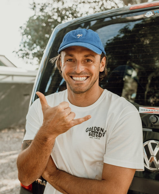Wearing a Palm Cap | Pacific and a white Golden Breed t-shirt, a man stands smiling with a shaka hand sign in front of a black vehicle, radiating laid-back beach vibes.