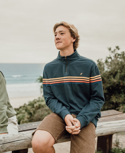 A young man in a blue pullover and Golden Breed’s Floyd Cord Walkshort in Wash Brown sits on a wooden bench near the beach, with trees and the ocean in the background.