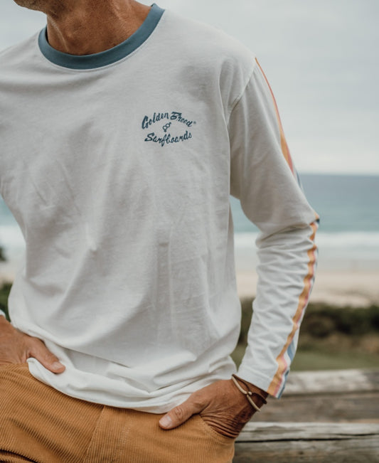 A person stands near the beach in an off-white Skater LS Tee by Golden Breed, featuring rainbow sleeve stripes and surfboard text. Made from 100% cotton and designed in Australia.