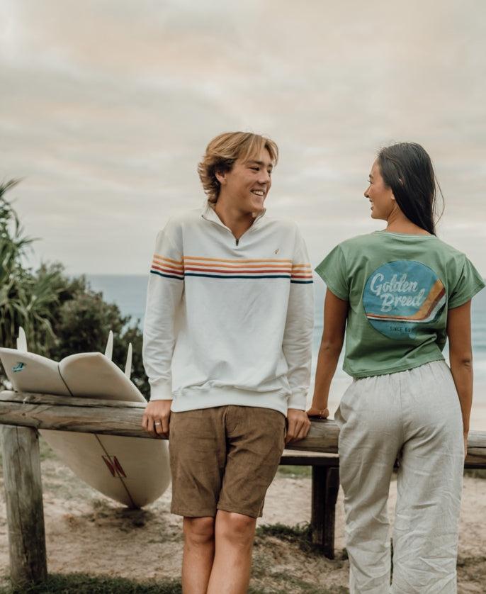 A young man and woman smile at each other by a bench with surfboards, wearing Golden Breeds Long Island Fleecy Top in Off White, surrounded by greenery and the ocean.