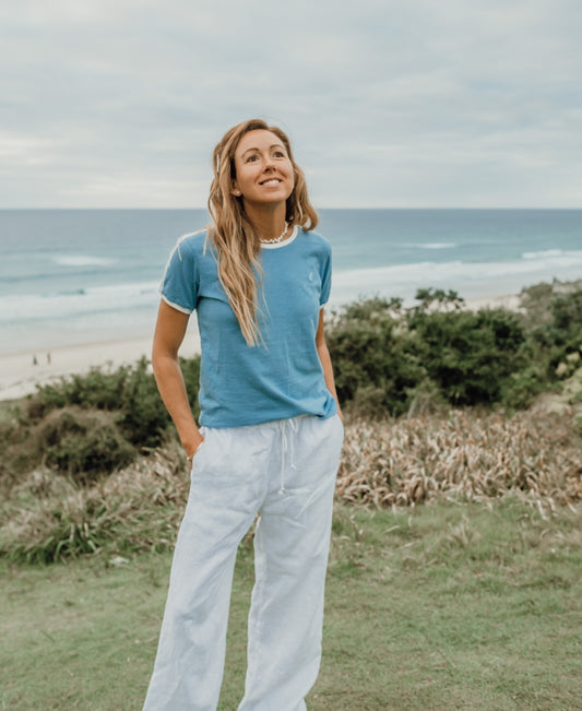 A woman stands on grass by the beach, wearing the Golden Breed Ringer Tee | Azure and white pants, with the ocean and a cloudy sky in the background.