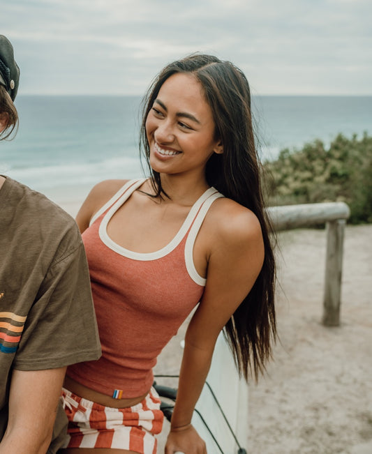A woman with long hair smiles by the beach, leaning on a railing and wearing the Golden Breed Flick Tank in Wash Red. Striped shorts complete her look as the ocean and sky create a scenic backdrop.
