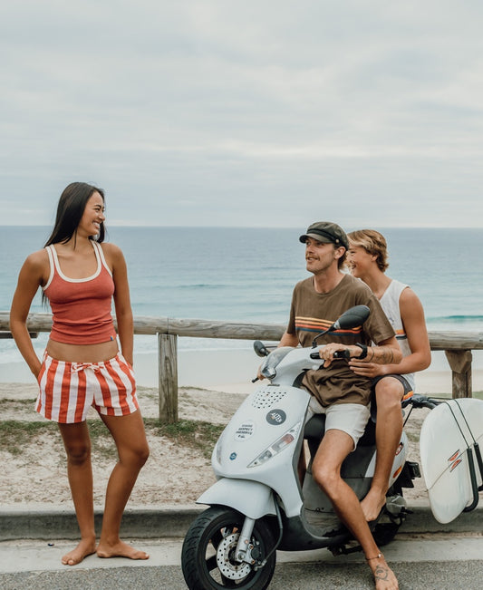 Two people wearing Golden Breeds Flick Tank in Wash Red sit on a scooter with a surfboard attached, while another stands nearby; theyre by the beach with the ocean in the background.