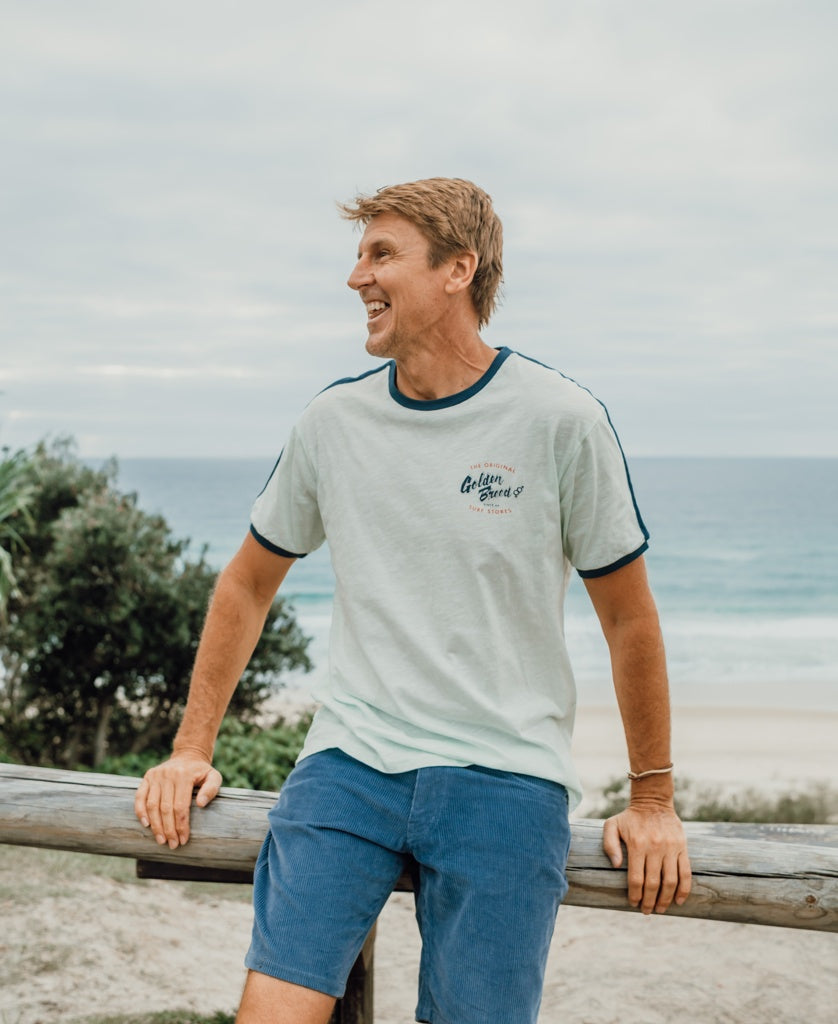 A man smiles as he leans on a wooden fence by the beach, wearing the Golden Breed Break Ringer Tee | Ice, designed in Australia from 100% cotton. The ocean serves as a scenic backdrop.