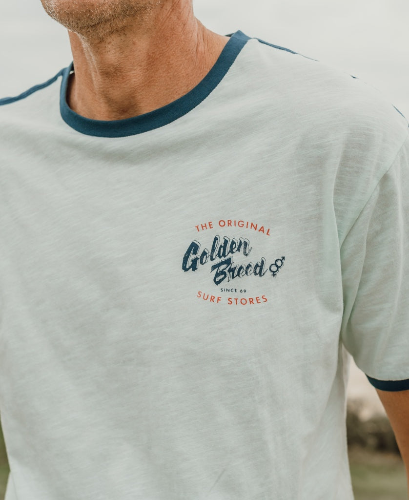 A person wears the Break Ringer Tee | Ice by Golden Breed, a light-colored tee with dark trim and a “Golden Breed Surf Stores” logo on the left chest. Designed in Australia.