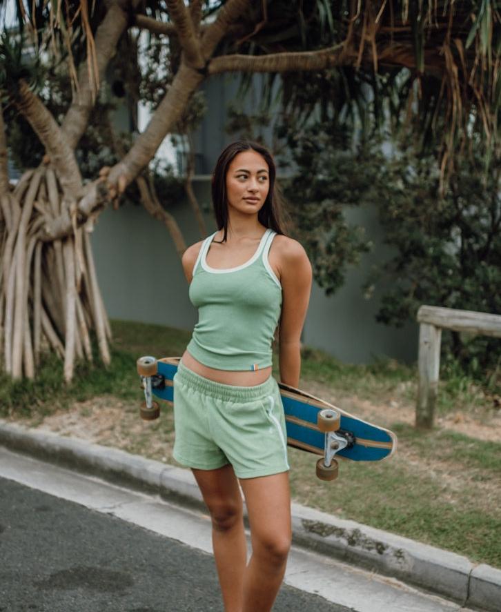A woman wearing the Golden Breed Flick Tank in Moss stands on a street holding a skateboard behind her back, with trees and a fence in the background.