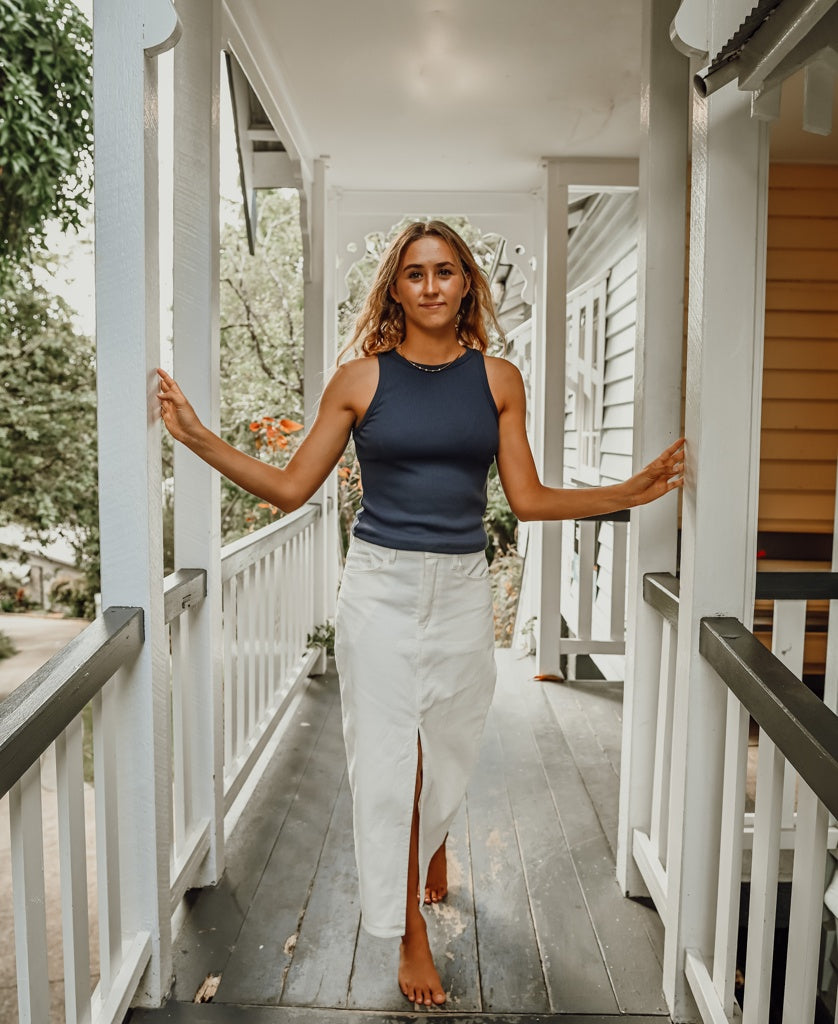 A woman with wavy hair, barefoot on a porch with white railings, wears the Golden Breed Nellie Maxi in Off White and a navy sleeveless top.
