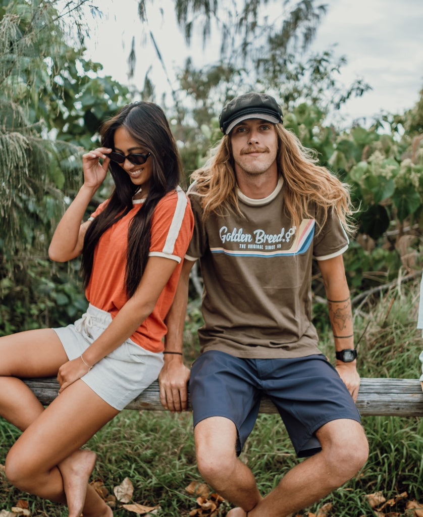 A man in a cap, shorts, and the Golden Breed Retro Ringer Tee in Wash Brown sits outdoors on a wooden rail next to a woman in sunglasses, red shirt, and shorts. Lush green foliage forms the backdrop. 70’s vibes abound.