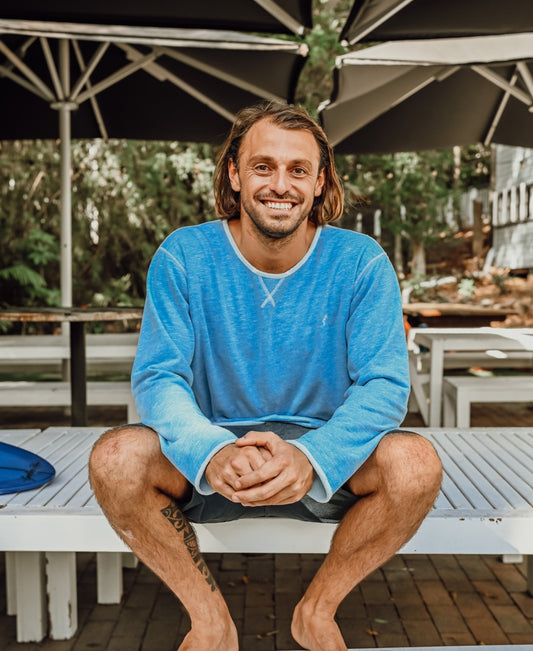 A man with long hair sits barefoot on a white bench outdoors, smiling and wearing the Golden Breed Classic Rag Top in Sky Marle. Large umbrellas and trees in the background complete his relaxed, retro surf style.