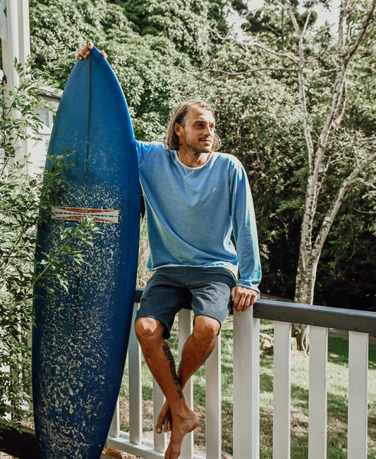 A man in the Golden Breed Classic Rag Top | Sky Marle shirt and shorts sits on a porch railing with a blue surfboard, showcasing retro surf style against a backdrop of trees and greenery.
